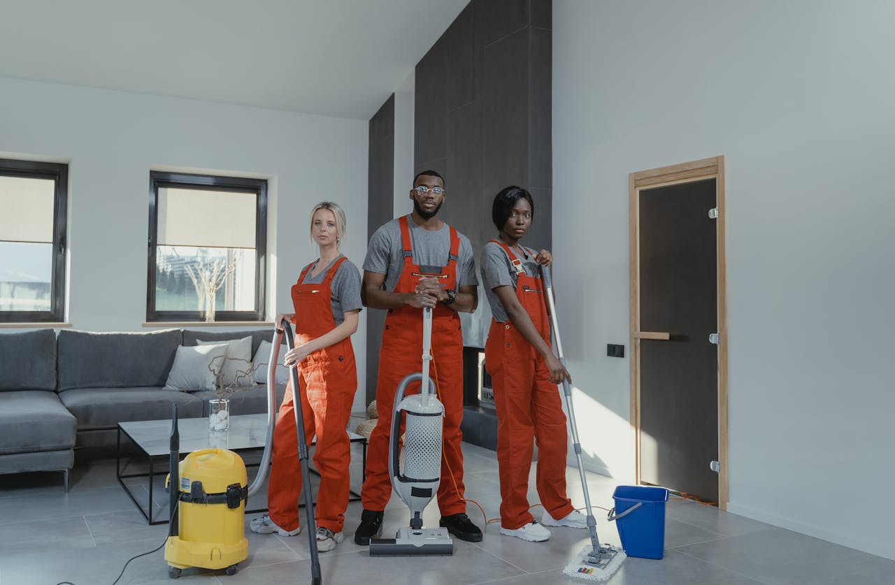 gallery-02 Diverse cleaning team in red uniforms ready to sanitize a modern living room with vacuum and mop.